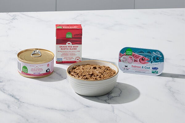 Image of a can of Open Farm wet cat food, a red container of wet cat food and a tin can of sardines for cats on a white countertop with a white bowl filled with wet cat food